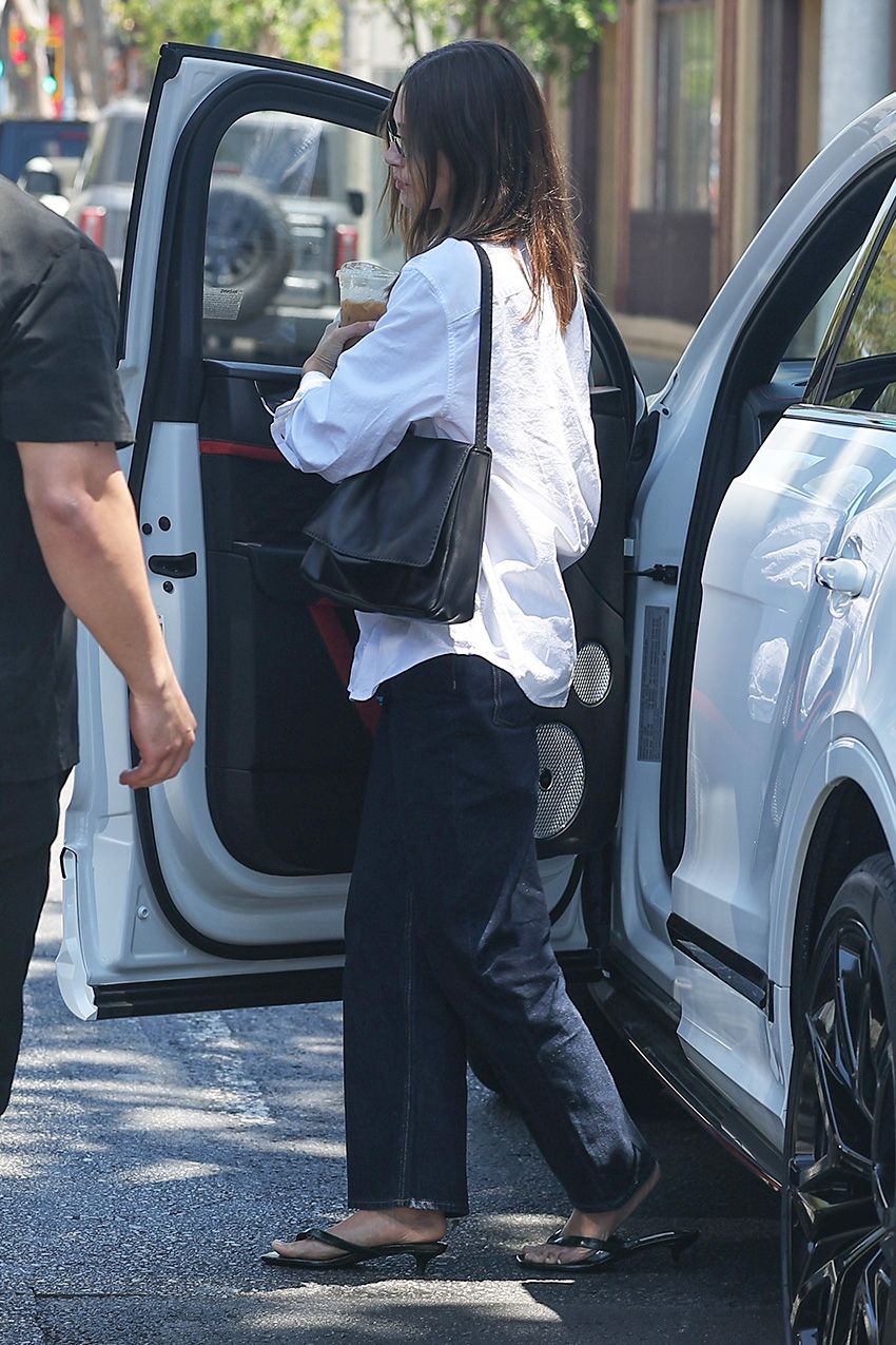 Hailey Bieber wearing a white button-down shirt with low-rise dark-wash jeans, kitten-heel thong sandals, and The Row shoulder flap bag