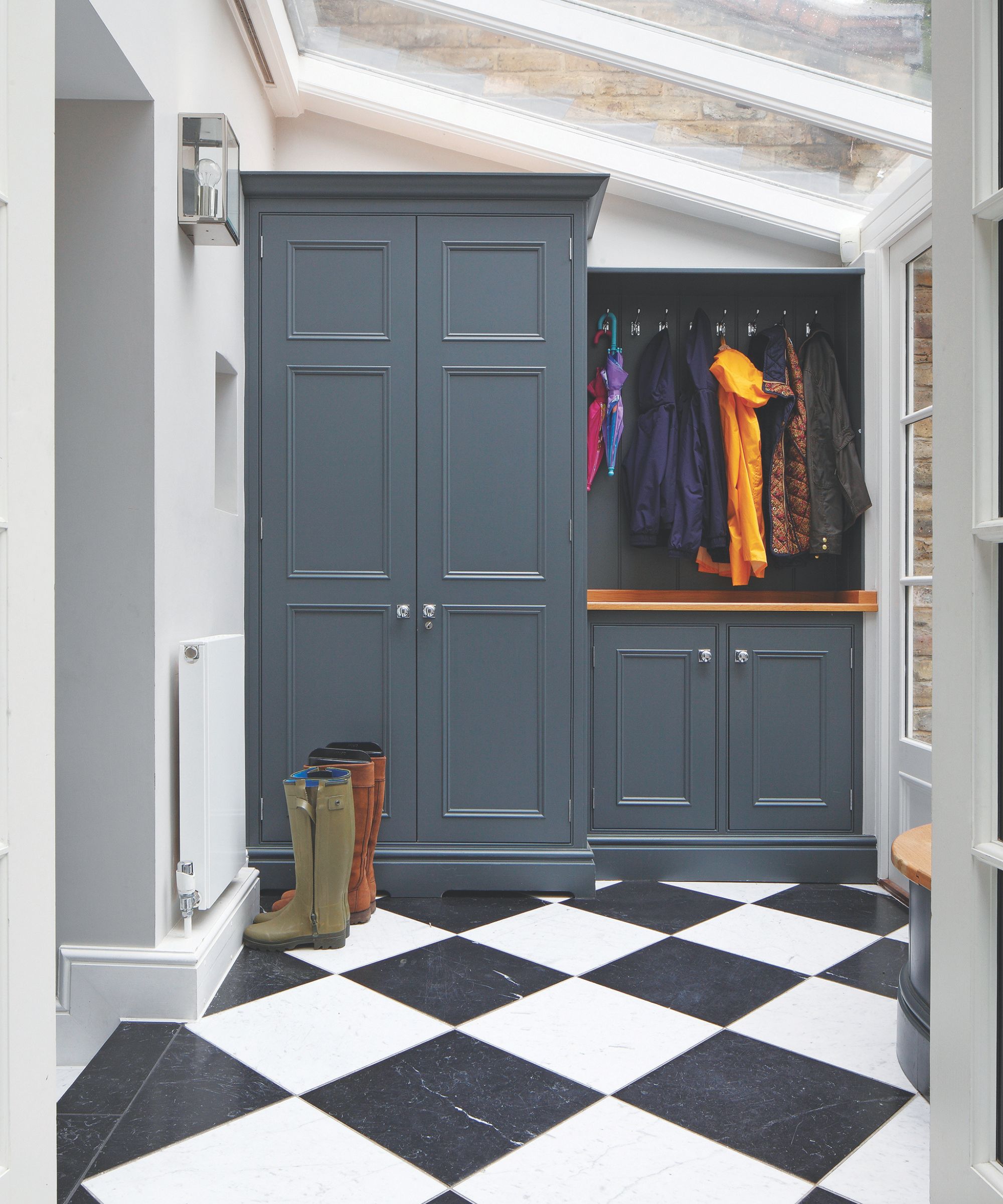 A bright mudroom with skylight and large windows, blue cabinets, black and white floor tiles, and wellies and jackets.