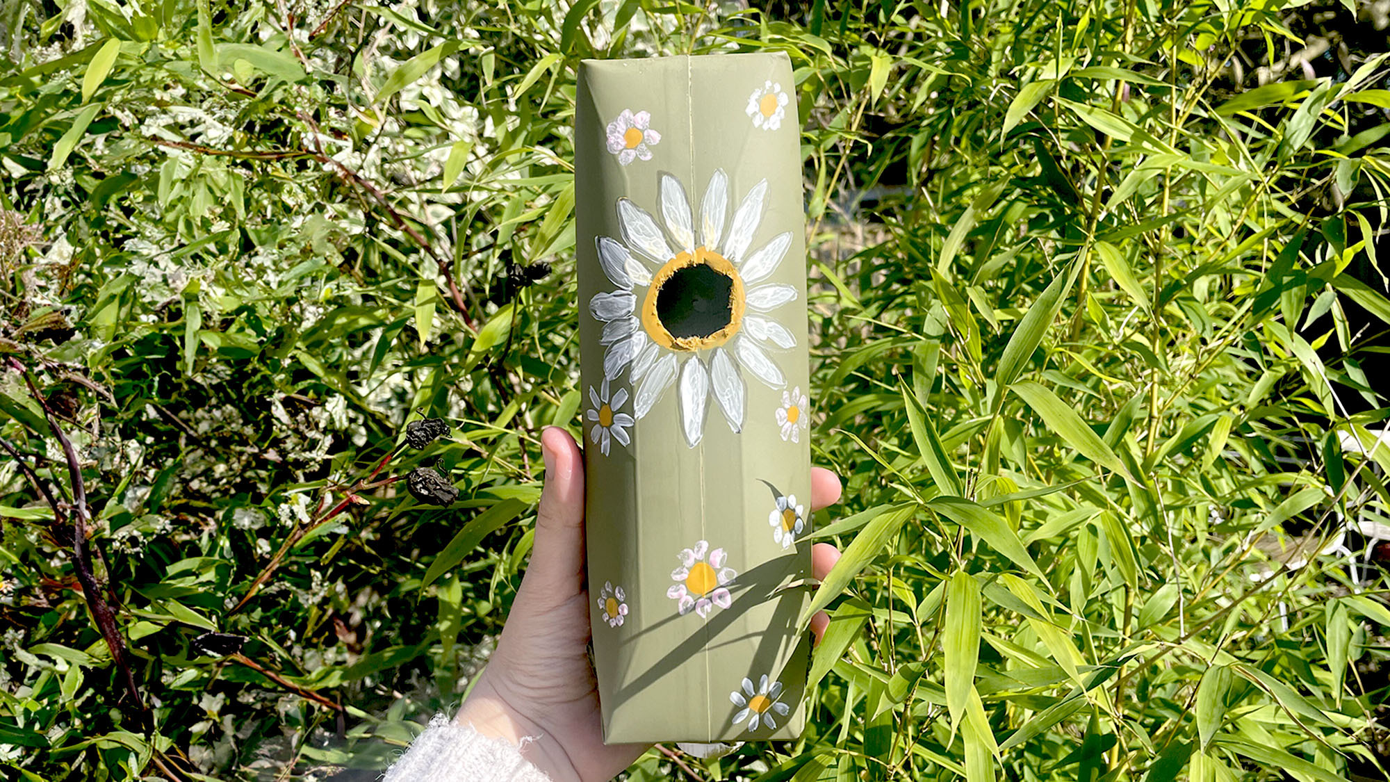 A homemade bird box created from a juice carton (painted green) held up by woman's hand against bamboo