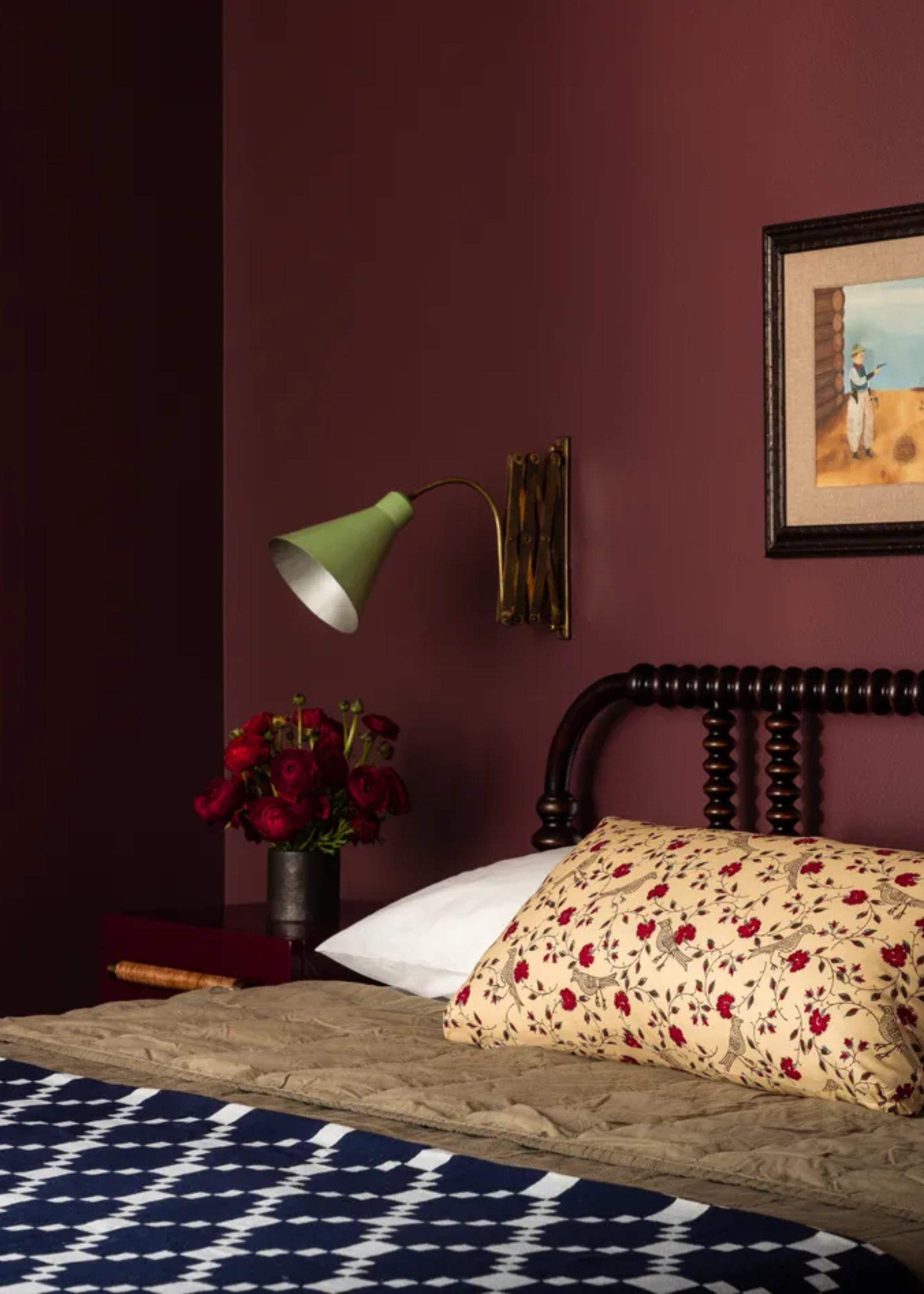 A burgundy bedroom with a framed painting, an extendable wall sconce, a side table with a vase of roses, a bobbin-style headboard, patterned pillow, and a patterned throw