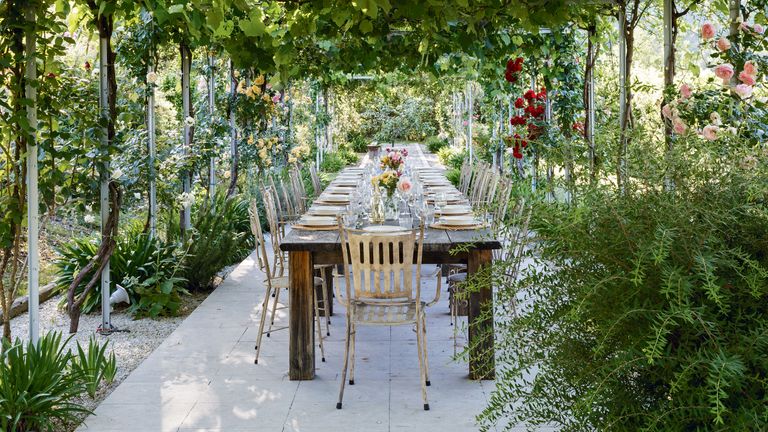 An example of garden shade ideas showing an outdoor dining area with a long wooden table surrounded by shrubbery