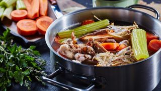 cooked chicken stock with vegetables and aromatic herbs in a stockpot and in a mug at the background, ingredients on a stone kitchen worktop, horizontal view, close-up