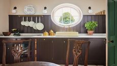 Rustic kitchen featuring dark brown wood paneling, white marble countertops, a cream-colored sink under a large oval window, and wooden dining chairs in the foreground