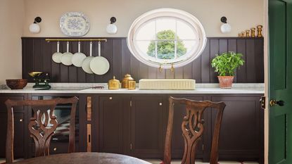 Rustic kitchen featuring dark brown wood paneling, white marble countertops, a cream-colored sink under a large oval window, and wooden dining chairs in the foreground