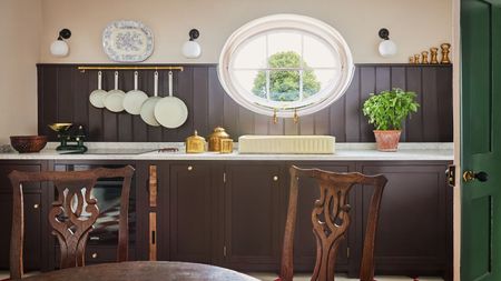 Rustic kitchen featuring dark brown wood paneling, white marble countertops, a cream-colored sink under a large oval window, and wooden dining chairs in the foreground
