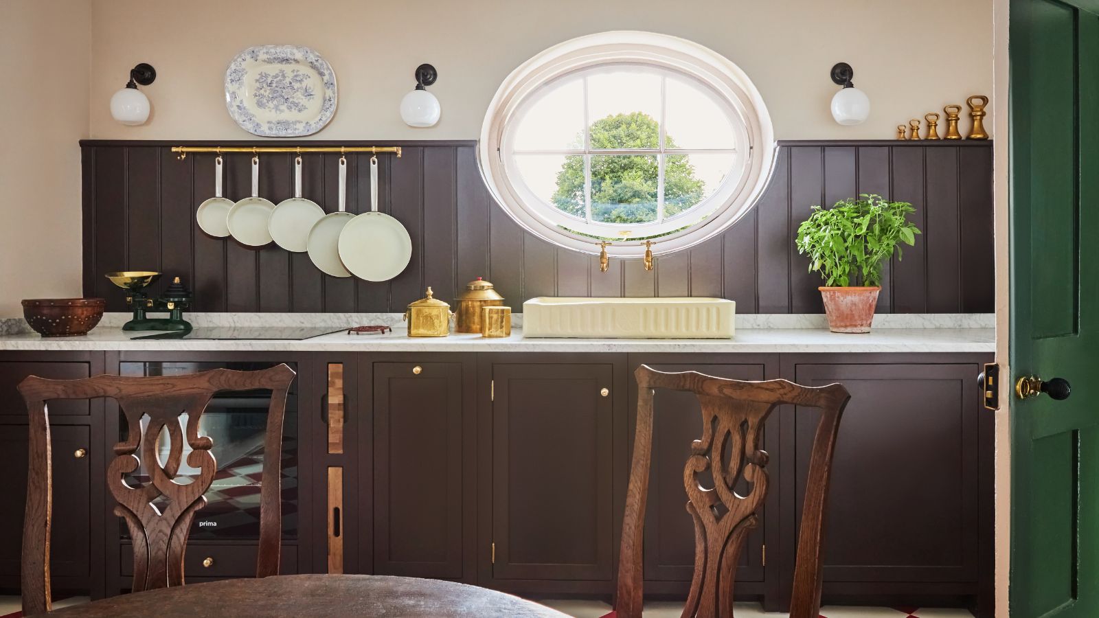 Rustic kitchen featuring dark brown wood paneling, white marble countertops, a cream-colored sink under a large oval window, and wooden dining chairs in the foreground