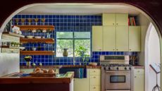 A large kitchen with a blue tile and white grout backsplash, mint green cabinets, wood floating shelves, in-built stove and oven, and a stand with a cookbook
