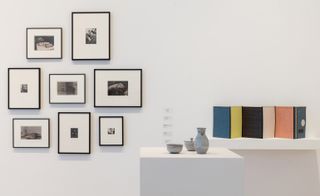 A white wall display of black frames (in different sizes) featuring black and white images. On the right of this is a white floating block shelf and in front is a white podium dressed with an open ceramic bowl, a closed ceramic bowl and a ceramic jug
