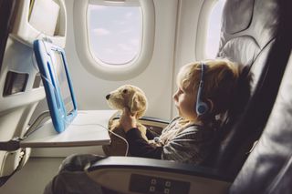 young child sitting in a window seat on a plane, wearing headphones and watching programmes on a tablet while holding a cuddly toy