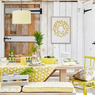 A rustic dining area with white wooden wall panels, a wooden table and chairs with yellow cushions and patterned pendant light