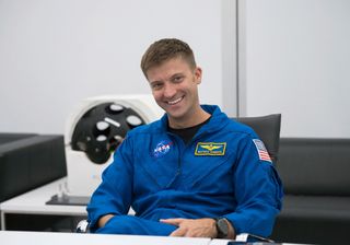 a man wearing a blue jumpsuit sits in a white room with black furniture. He is smiling.