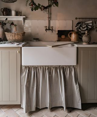 A farmhouse-style kitchen sink with a white ceramic apron-front basin, framed by cream cabinetry with a striped fabric curtain below. The backdrop includes square white tiles and a brass wall-mounted tap.