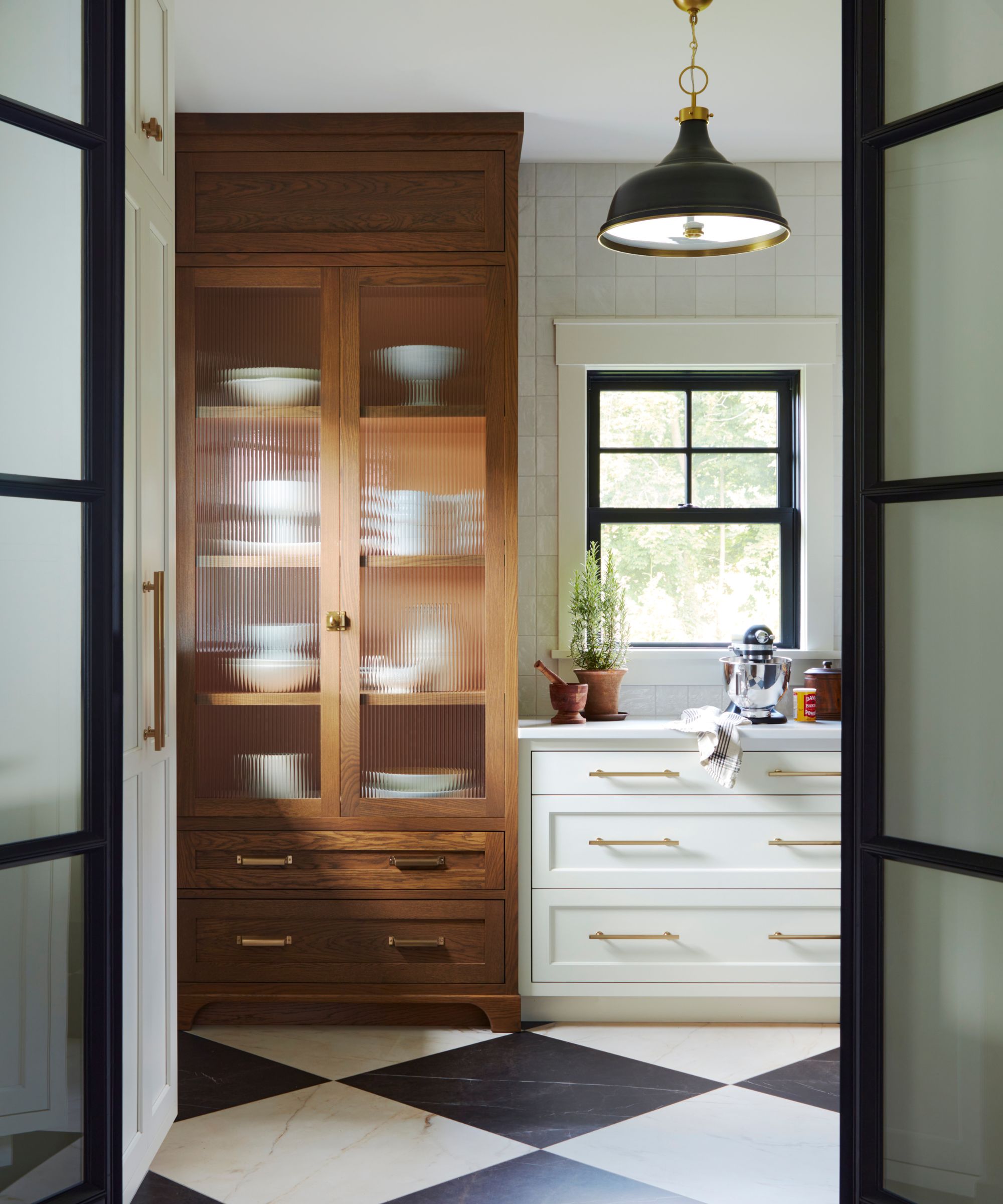 wooden and white kitchen cabinetry in a back kitchen seen through black metal and glass doors with a black and white checkerboard tiled floor