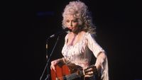 Dolly Parton speaks from the stage as she stands with her acoustic guitar, 1978.