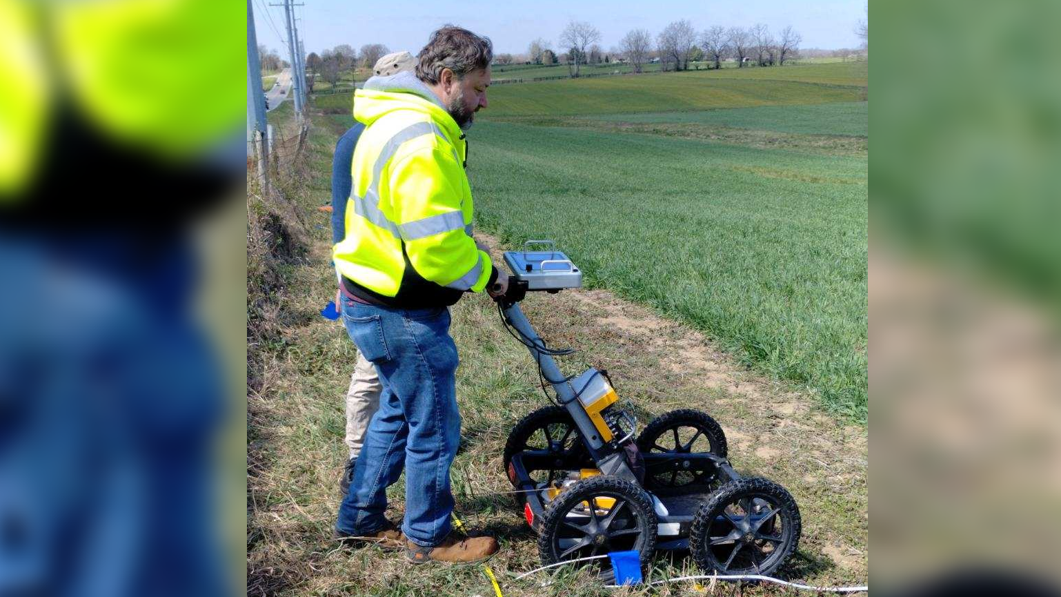 A man in a blaze yellow vest pushes a contraption that looks like a vacuum with four wheels in a field.
