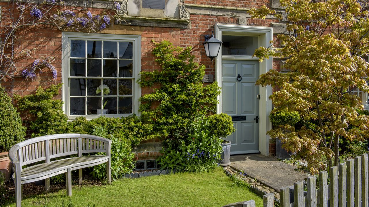 Exterior of a former school house, a Victorian village house built in red brick, and garden bench by the front door.