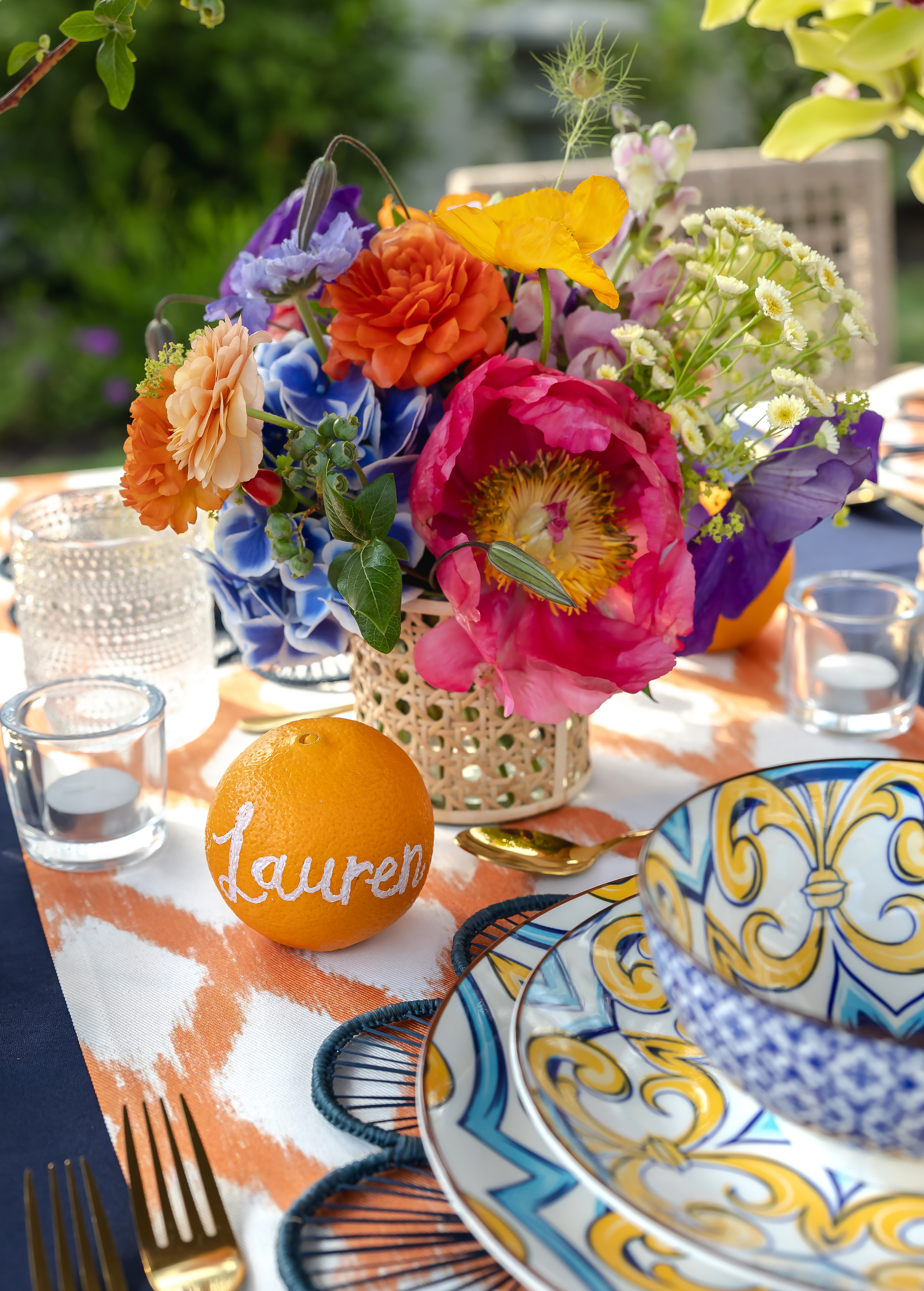 An orange printed table cloth with blue and yellow patterned plates, and a bright floral bouquet, with decorative glass tea lights.