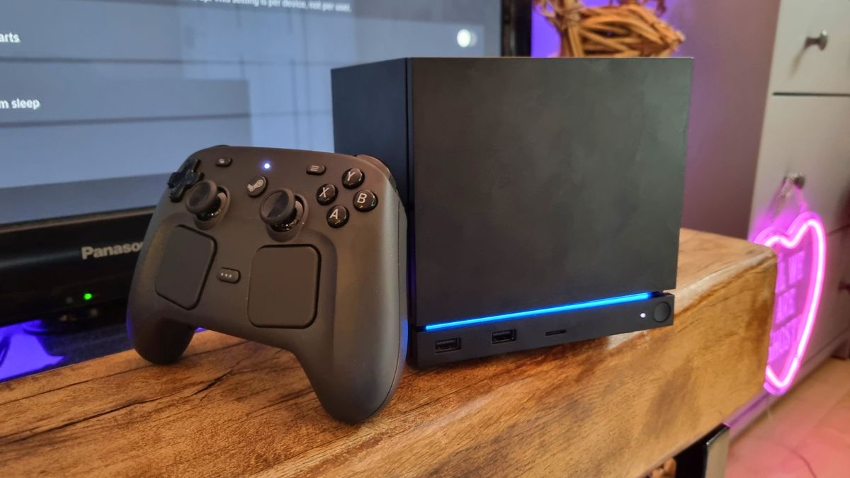 A Steam Machine in front of a TV, with the Steam Controller resting against it and a pink neon sign in the background