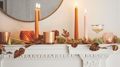White Autumnal mantelpiece with lit orange taper candles and leaf garland and circular mirror
