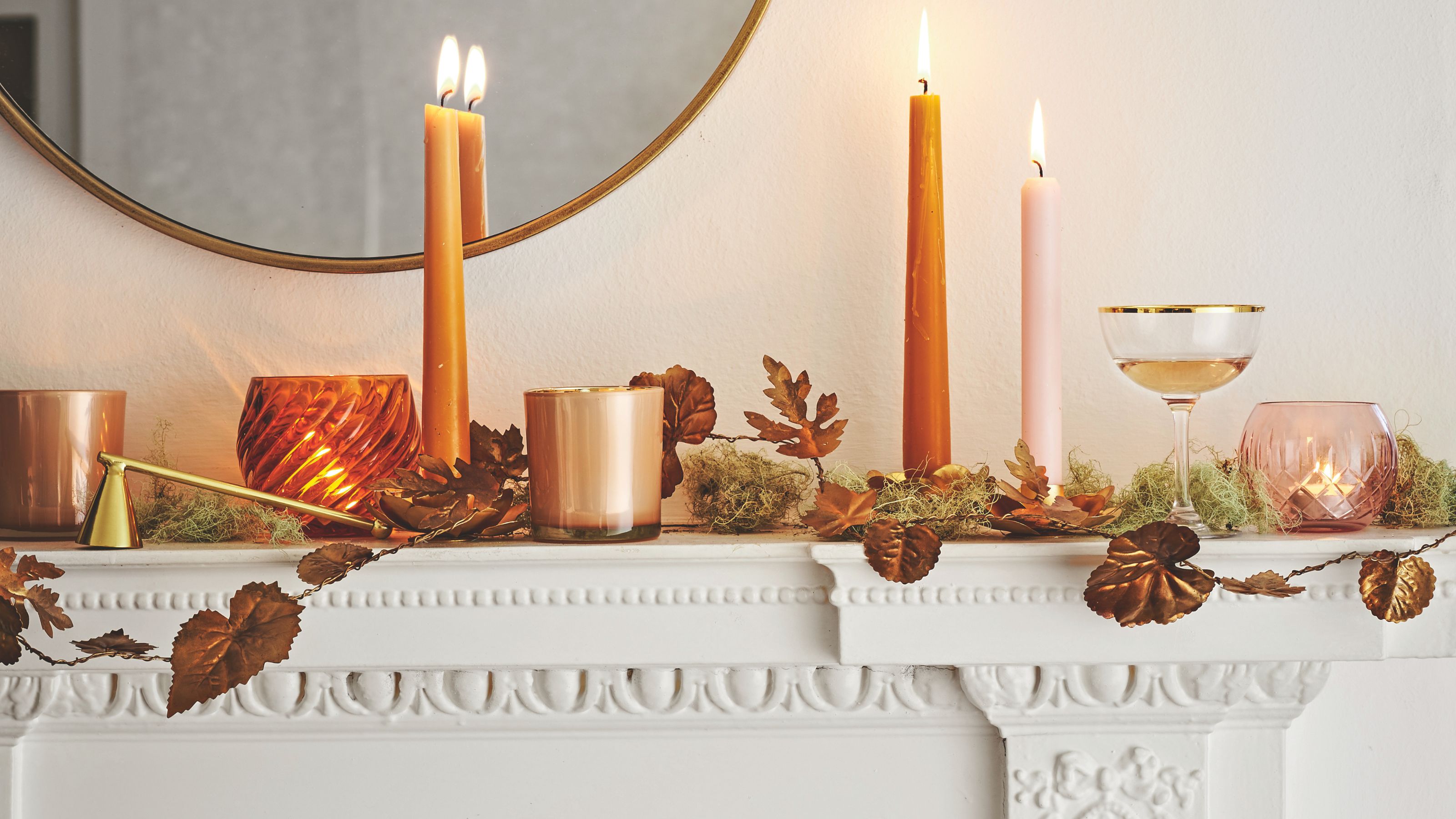 White Autumnal mantelpiece with lit orange taper candles and leaf garland and circular mirror