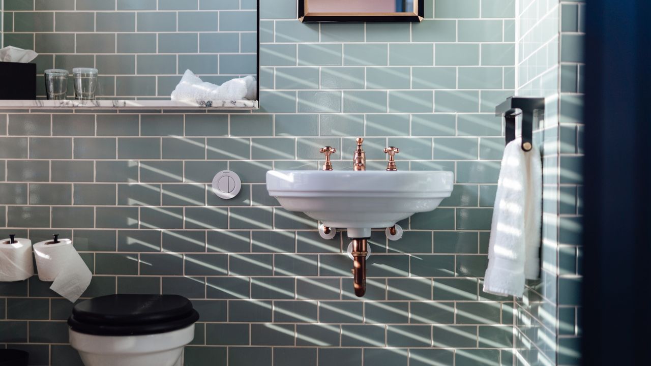 Sunlight coming through from the window in a stylish bathroom with light green brick-look wall. Includes white toilet, with black lid, white sink with gold taps, and hanging fluffy-white towel