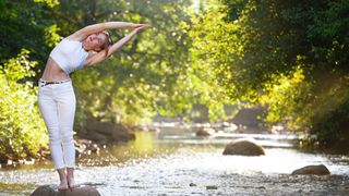 A woman does a standing side stretch on a rock in a river