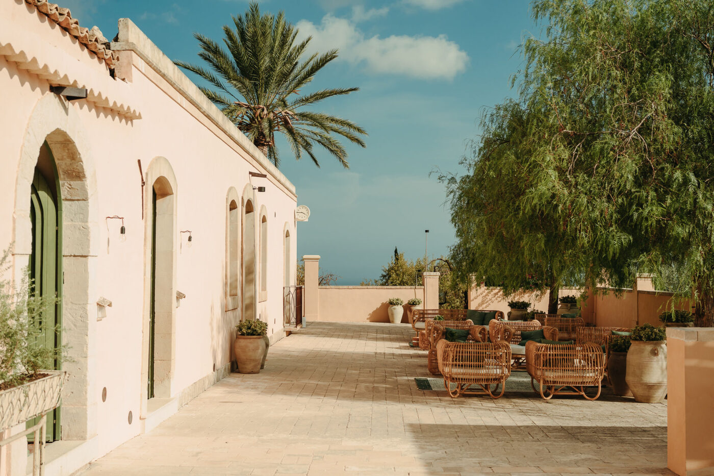 A sprawling Sicilian hotel rises within a dense, sun-kissed olive grove, boasting red and white tiling, rattan roofs, and white-washed walls.