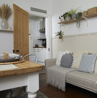 Grey sofa with neutral colored pillows next to a wooden dining room table with a wall-mounted shelf topped with plant-décor and woven accessories