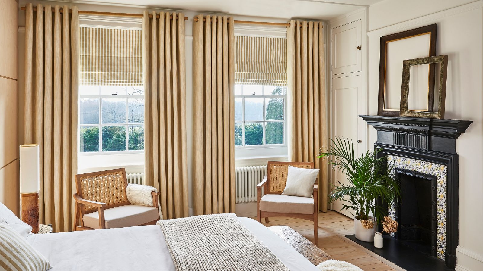 A neutral bedroom with beige curtains and blinds. Two rattan arm chairs face the bed, and a black fireplace opposite. 