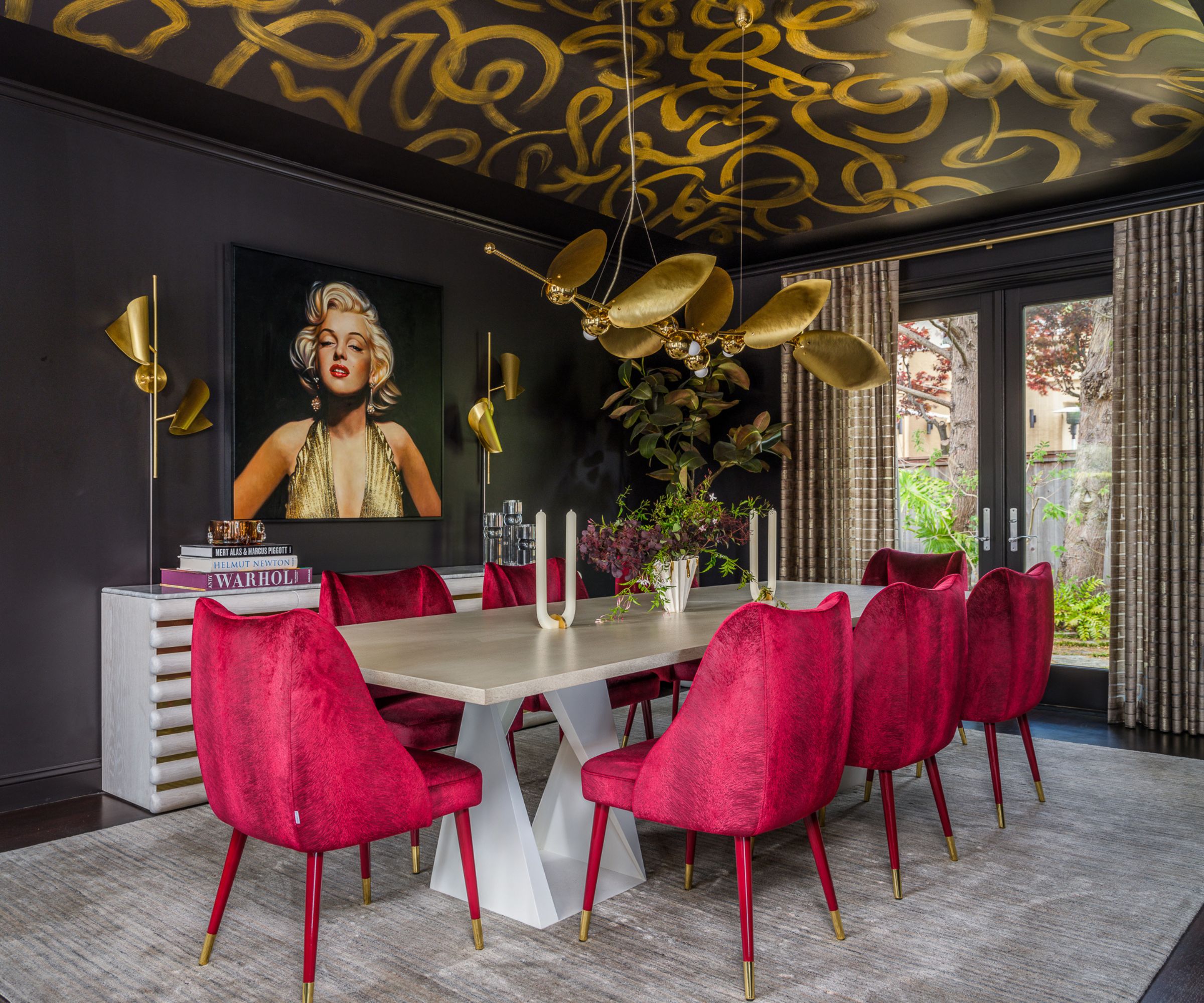 Bold dining room with black painted walls, patterned wallpapered ceiling, white dining table with 9 pink chairs, large gray rug underneath, sculptural gold pendant light, artwork hung behind.