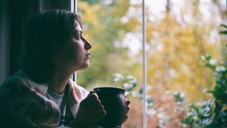 depressed woman sitting near window and looking at autumn landscape outside in rainy day.