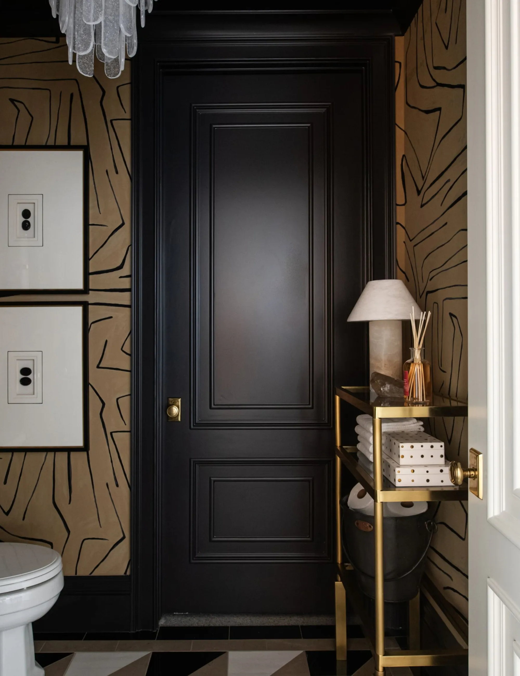 A moody bathroom with black millwork, a gold and black wallpaper, and a gold console decorated with a reed diffuser and lamp