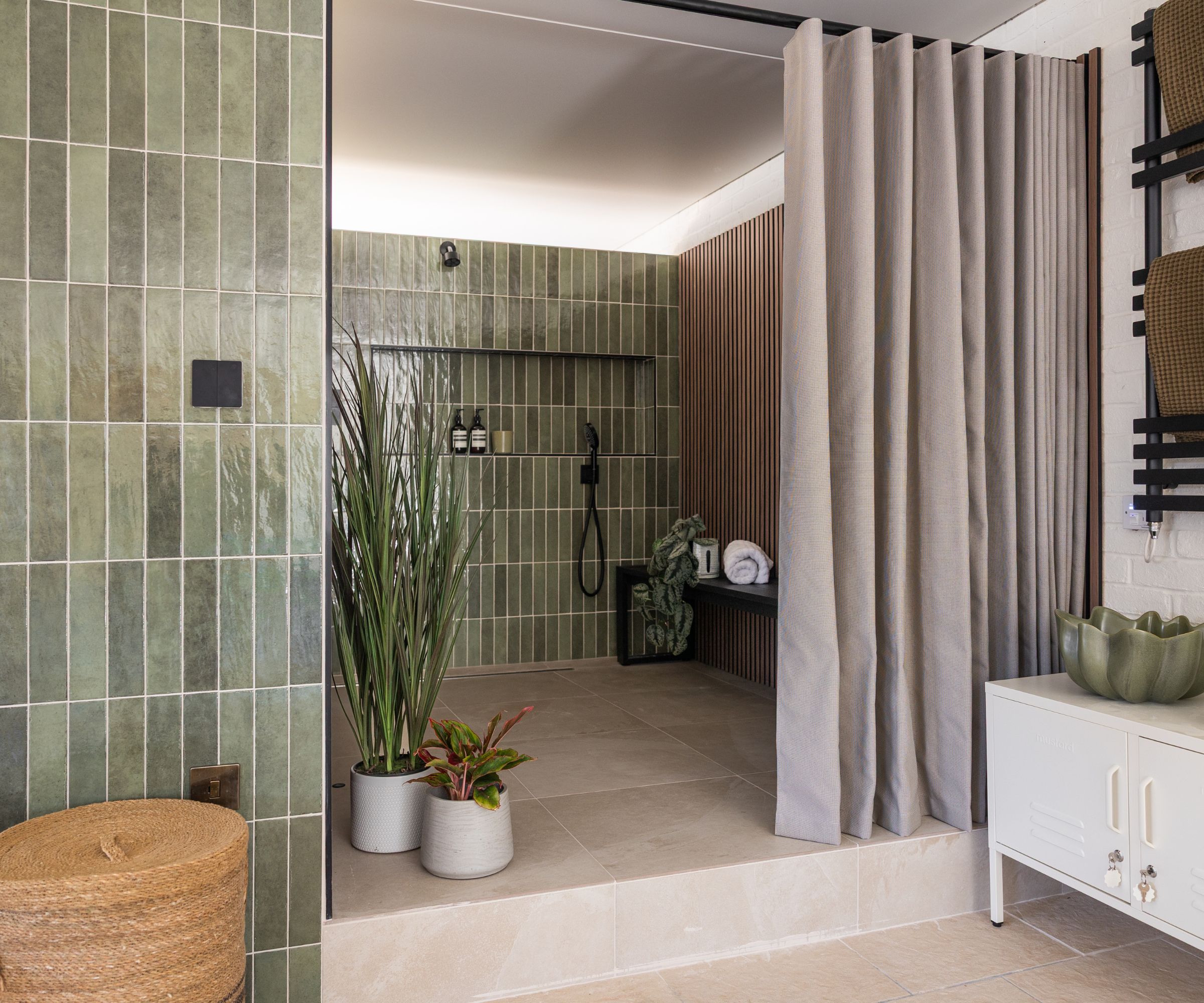A pool house changing room with green glazed vertical tiles, a curtained wet room with black shower fittings, wood slat panelling, potted plants and a black heated towel rail.