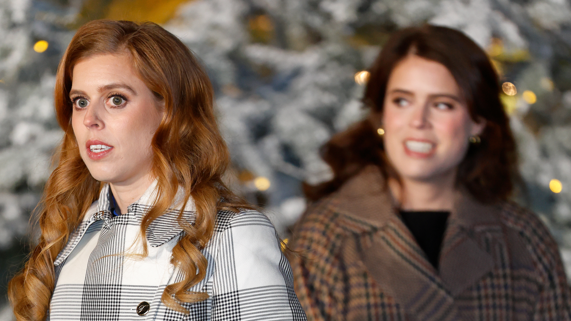 Princess Beatrice wearing a plaid coat and Princess Eugenie wearing a brown wrap coat in front of a flocked Christmas tree