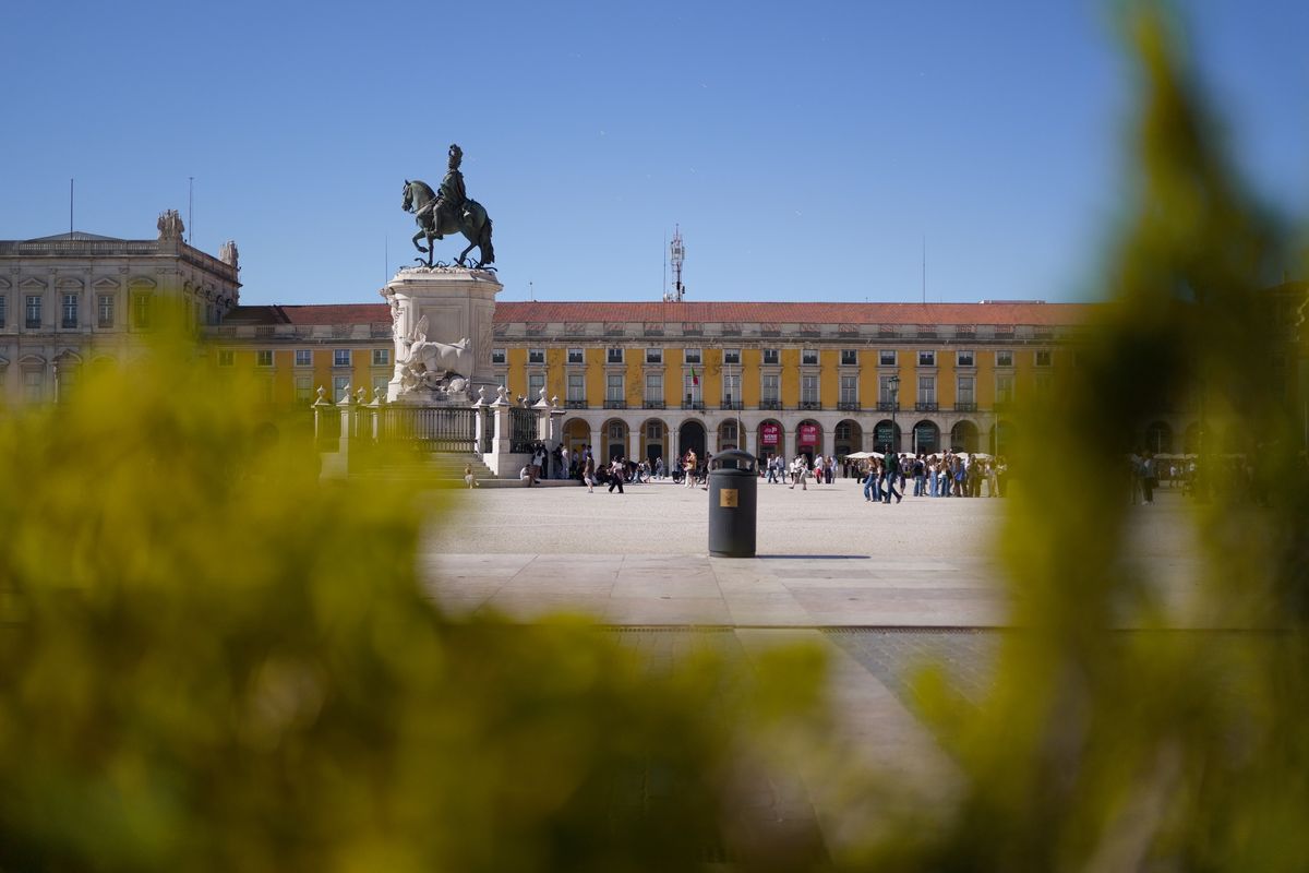 Equestrian statue in Pra&ccedil;a do Com&eacute;rcio framed through out-of-focus green foliage, with yellow historic buildings behind.
