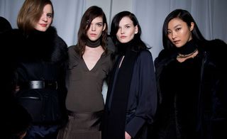Four female models wearing looks from the Ports 1961 collection. One model is wearing a dark blue belted jacket with black fur and dark blue bottoms. Next to her is a model wearing a brown V-neck top and brown skirt. The third model is wearing a dark blue top and black scarf. And the fourth model is wearing a black fur piece with a dark blue jacket over the top