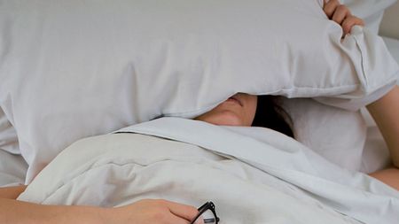 person lying on bed while covering face with pillow and holding eyeglasses