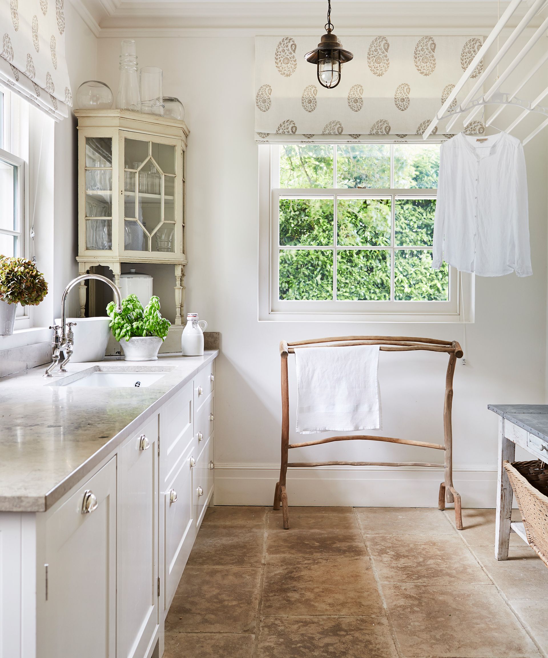 A light and airy kitchen with white cabinets and a stone floor in front of a large traditional window with decorative blinds