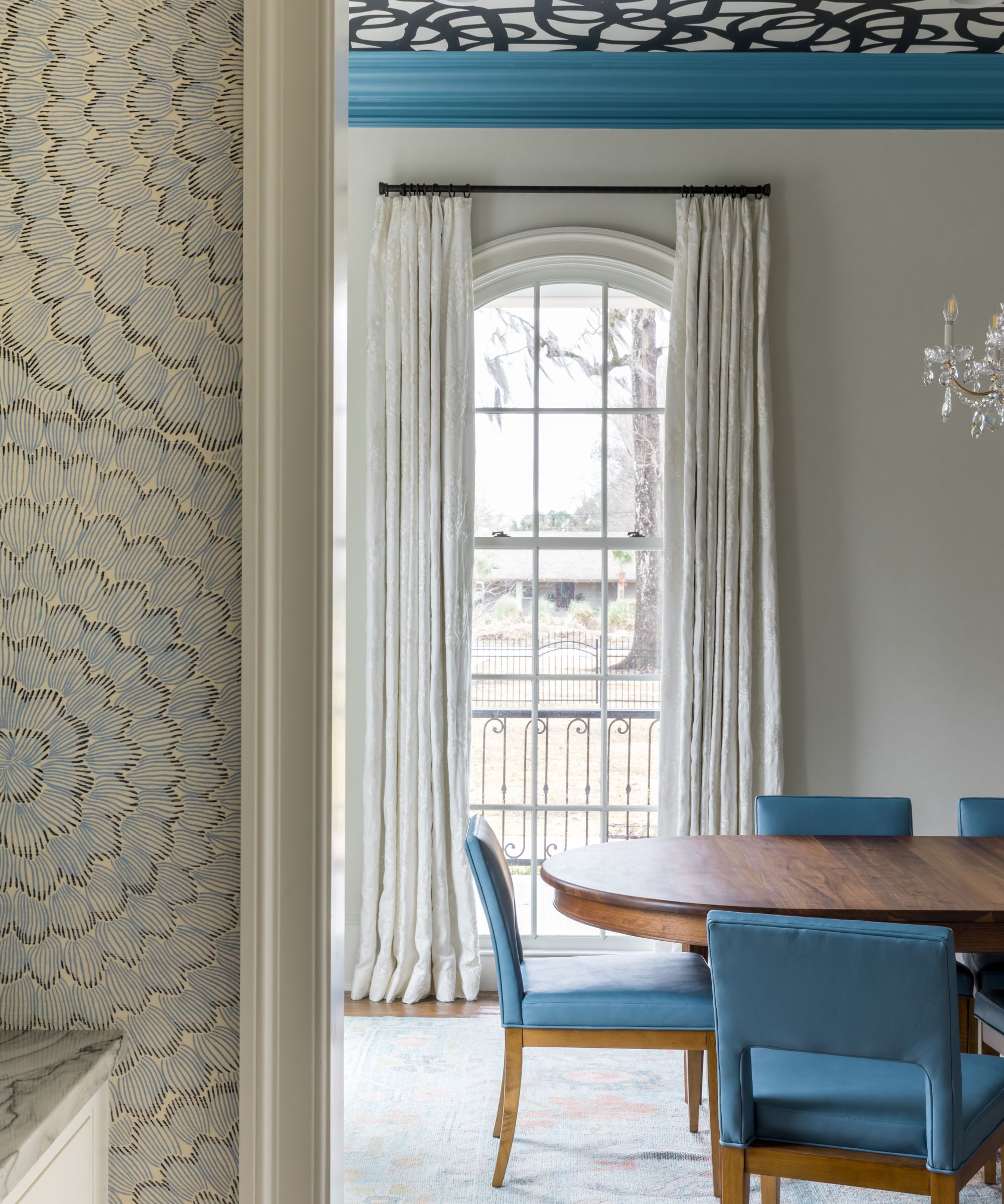 A dining room in a large room with a high ceiling. White walls, a tall arched window with white curtains, blue ceiling trim, and a black-and-white patterned wallpapered ceiling. A dark wood dining table with blue chairs.