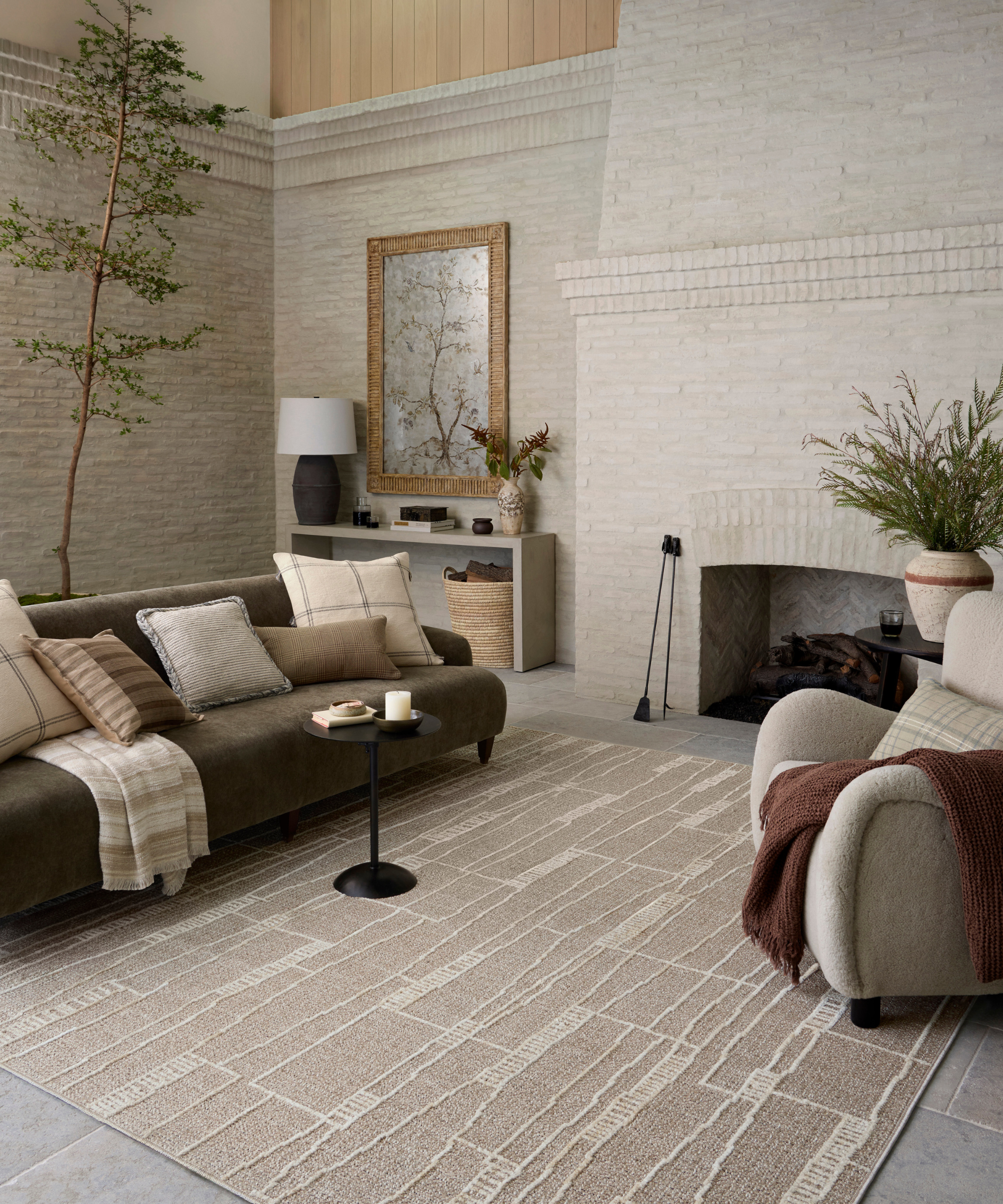 a living room with brick walls and a large brick hearth, a dark green couch with various pillows, a small black drink table, a hi-low pile rug in tan, and an accent chair on the right