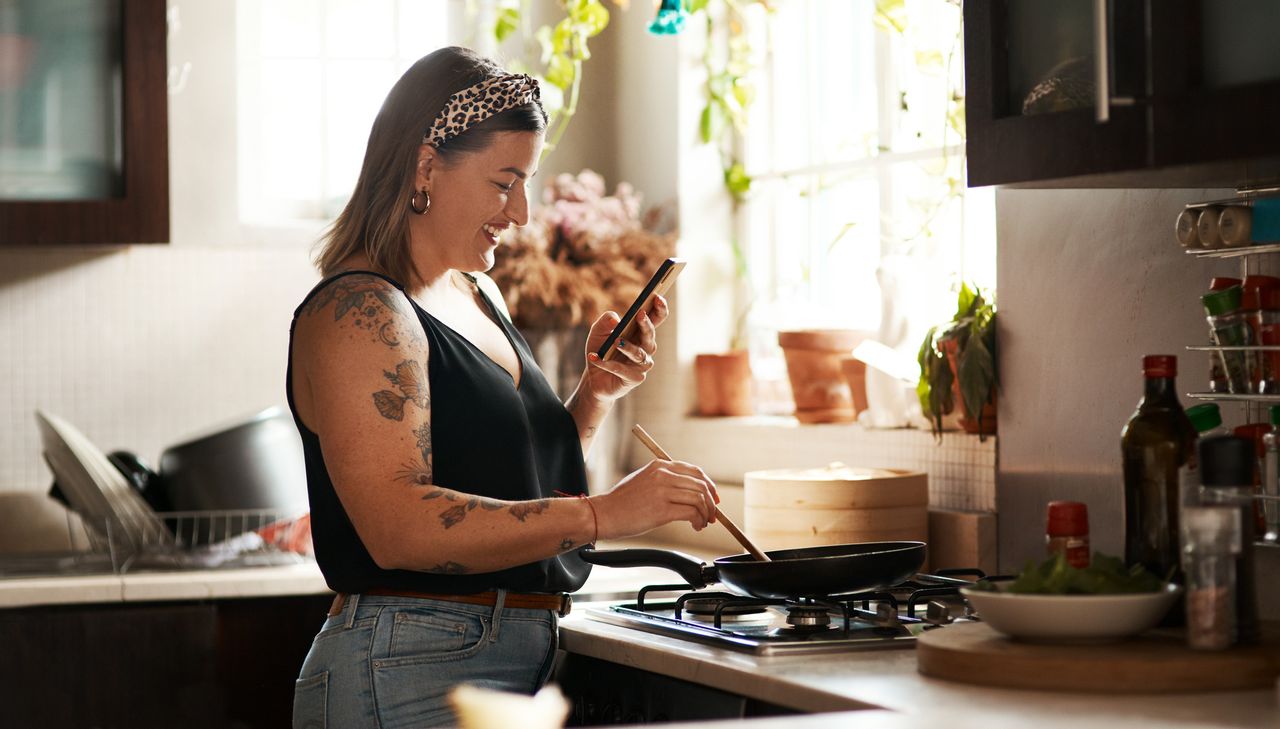 Woman stands at gas stove, using wooden spoon to stir food cooking in a frying pan, while looking at a smartphone in her other hand. She wears light blue jeans, a black sleeveless top and a leopard-print bandana headband