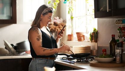 Woman stands at gas stove, using wooden spoon to stir food cooking in a frying pan, while looking at a smartphone in her other hand. She wears light blue jeans, a black sleeveless top and a leopard-print bandana headband