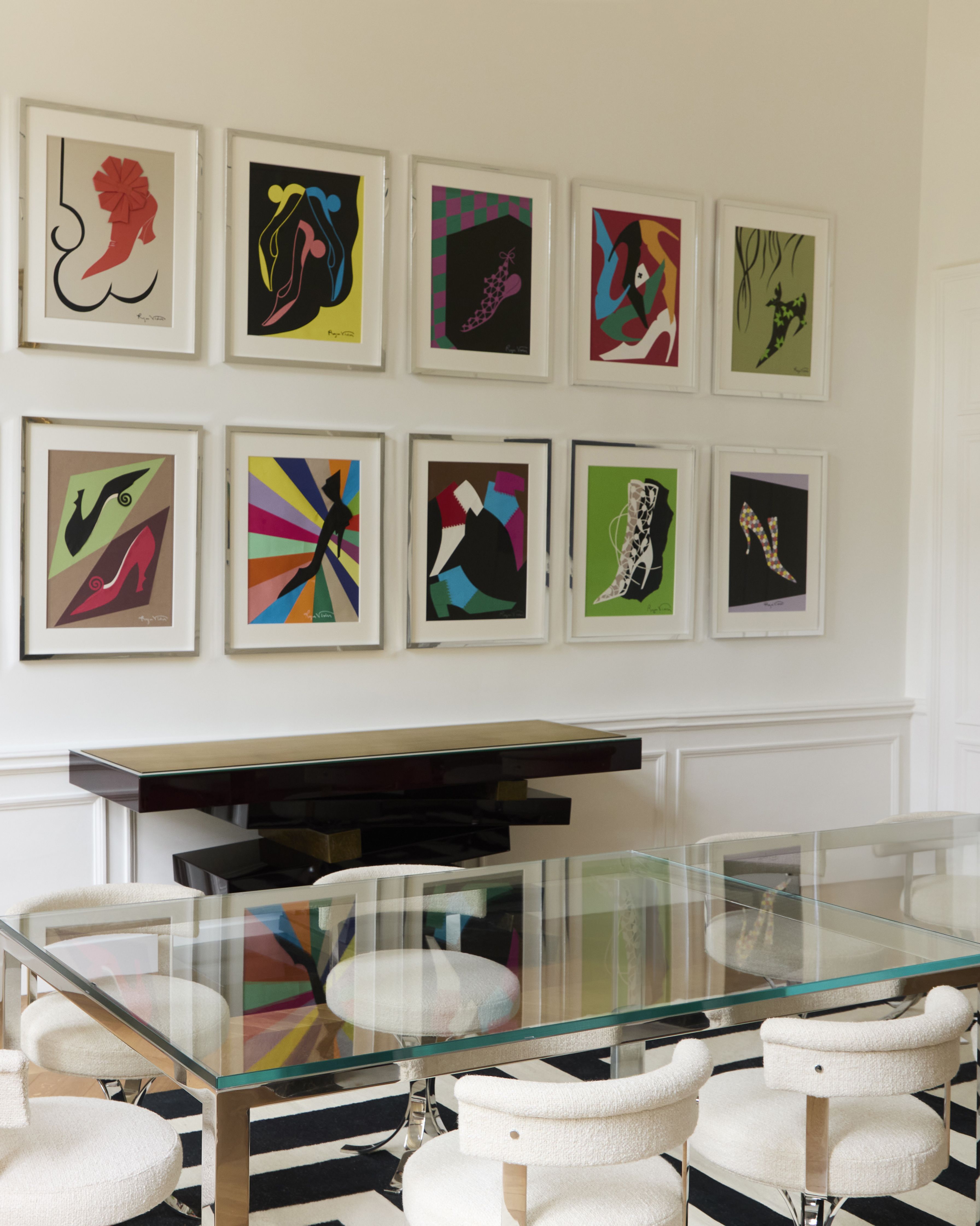A maximalist salon features a chrome and glass table with shearling and chrome matching chairs, an unadorned black cabinette with a sculptural, zig-zag structure, and, on top of it, a series of framed, colorful prints depicting shoes, framed in silver.