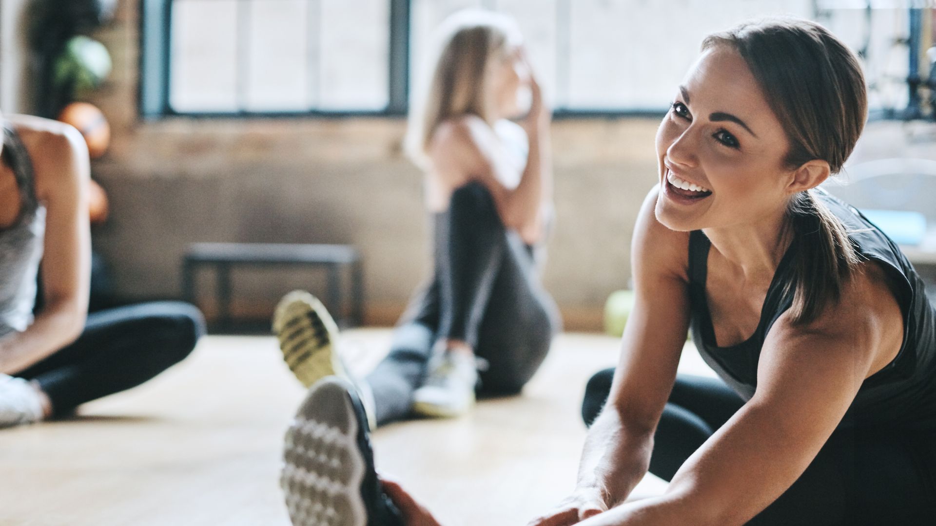 Woman stretching before step-up exercise in the gym, sitting with others on wooden floor