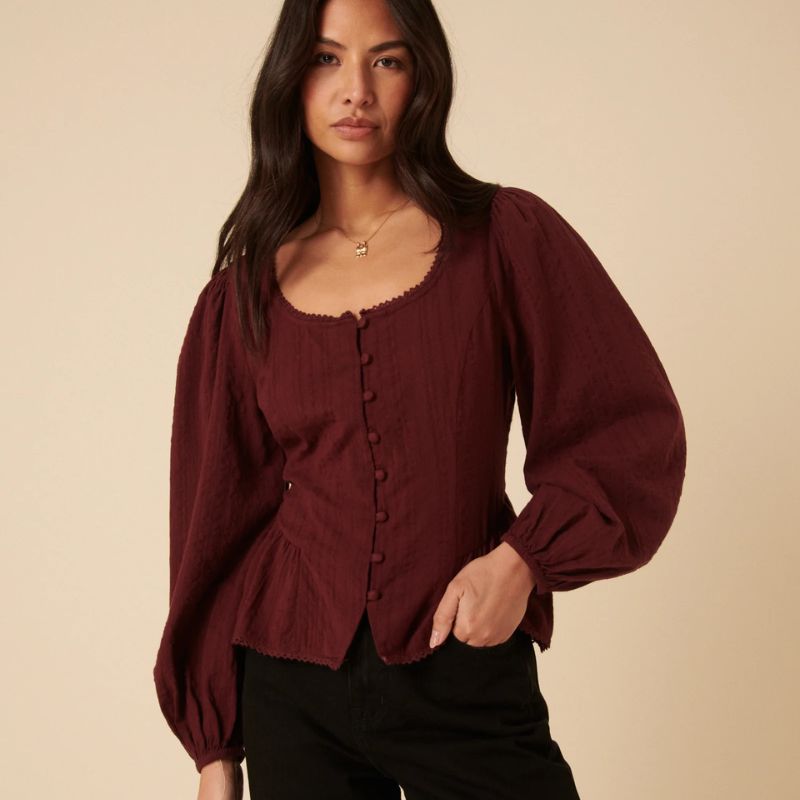 A model with dark hair wears a scooped neck burgundy blouse with black jeans.
