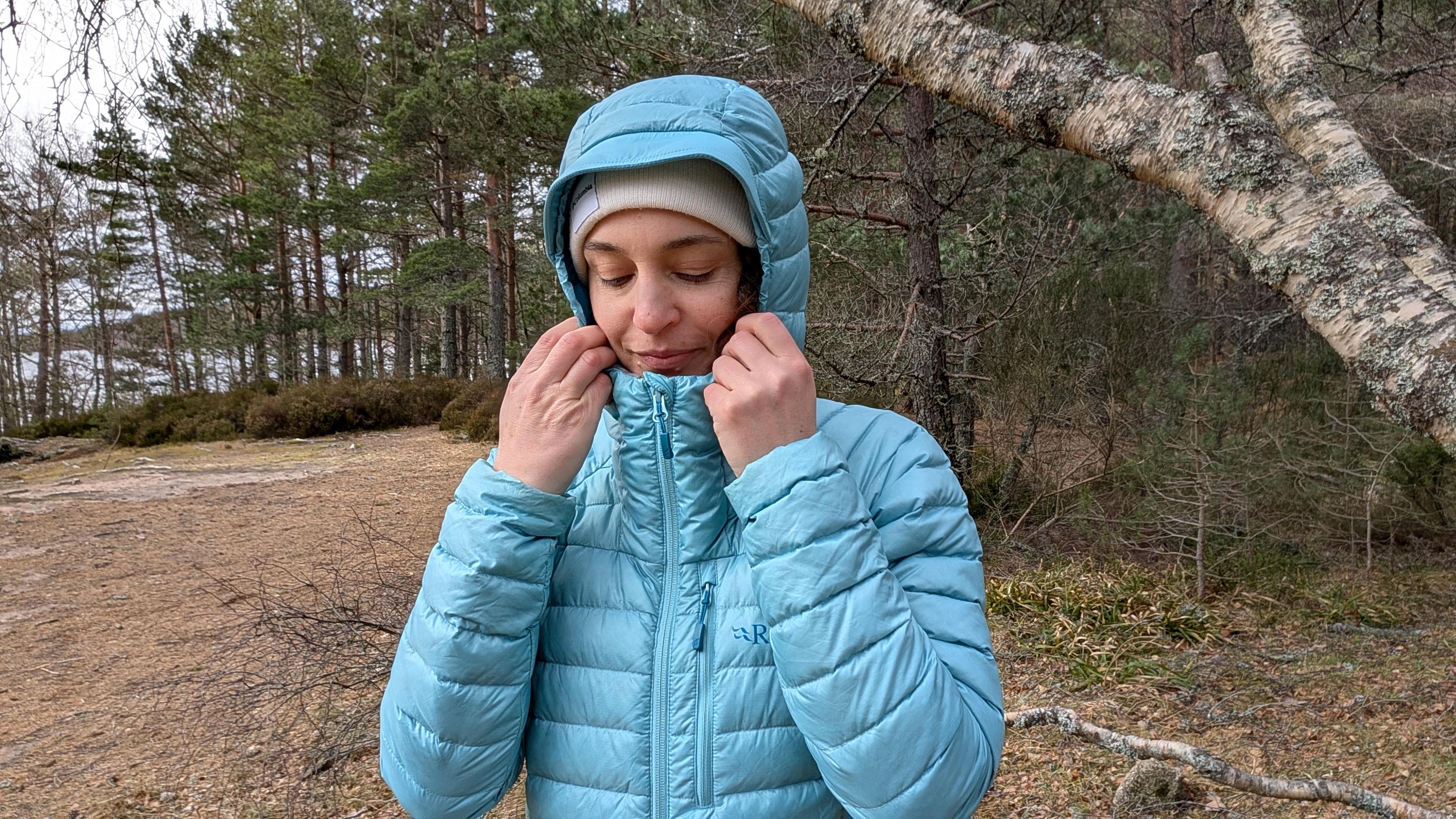 A woman on a beach near a lake wearing putting up the hood of her bright blue down jacket