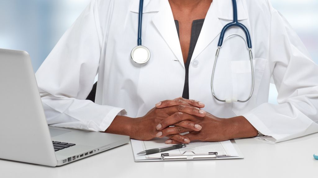 woman in lab coat with stethoscope around her neck and hands on the table in front of her 