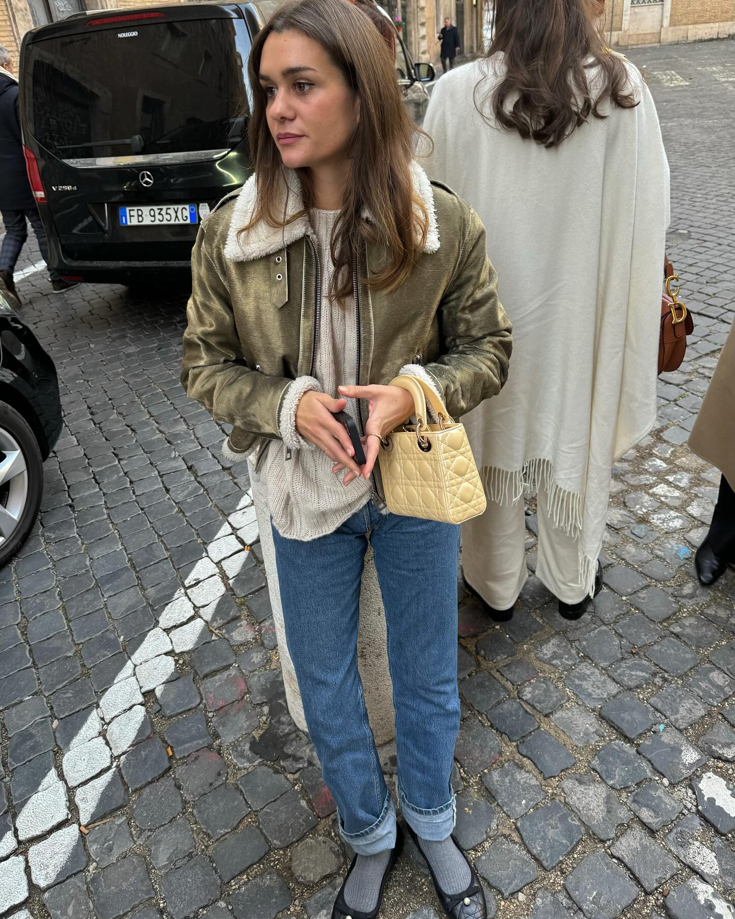 French fashion influencer poses on the cobblestone streets of Paris in a shearling lined jacket, neutral sweater, yellow mini Dior bag, cuffed jeans, gray socks, and black ballet flats