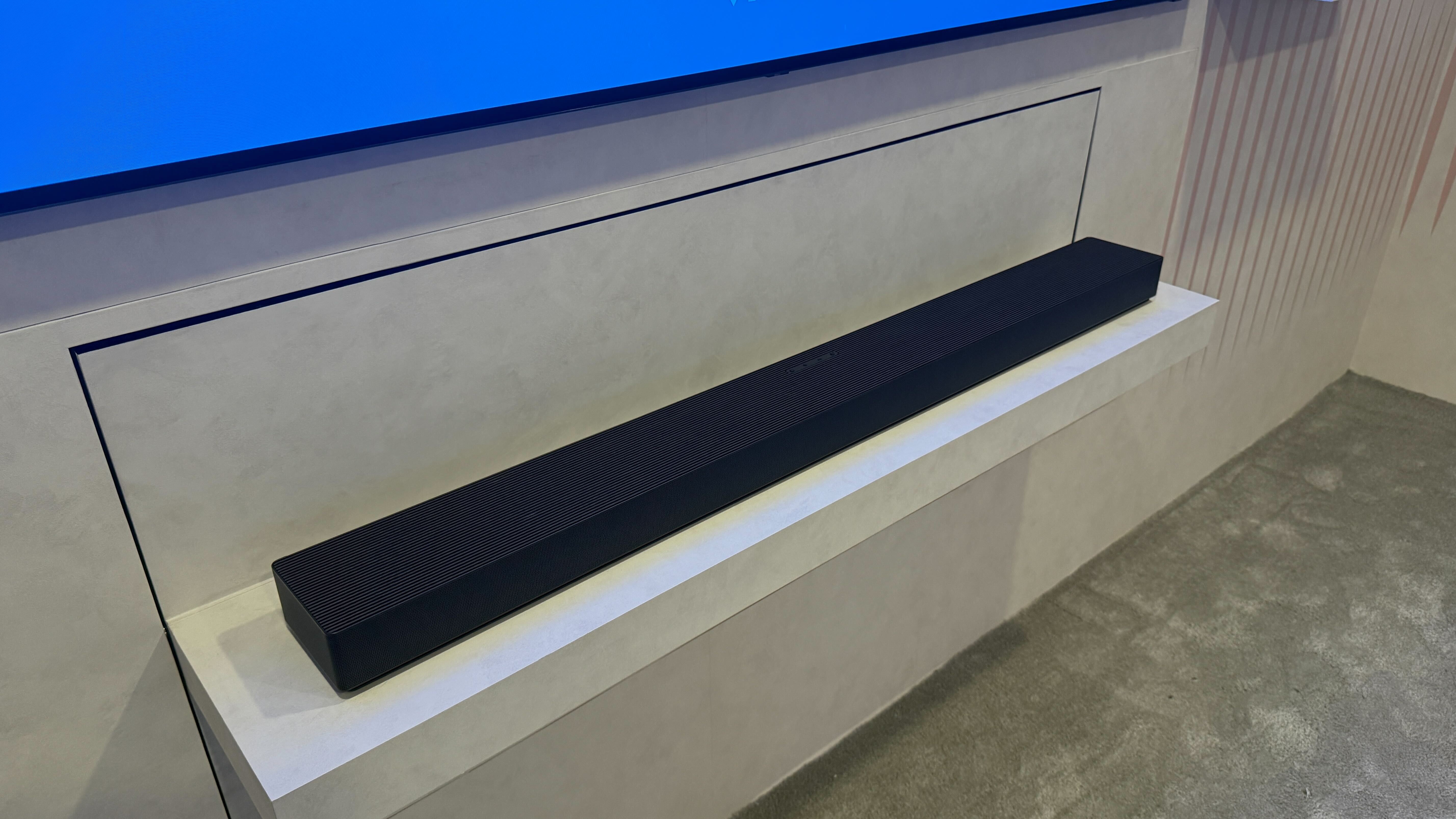 Samsung's HW-QS90H sitting below a wall-mounted TV on a white backdrop