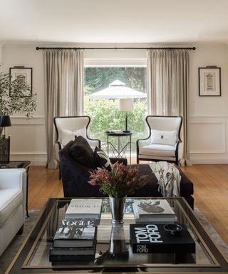 A family room with white walls, black and white accent chairs, off-white curtains and black and gold coffee table